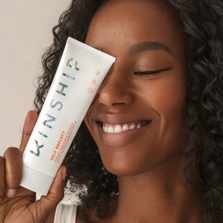 Smiling woman holding Kinship Self Reflect Probiotic Sunscreen SPF 32 tube close to her face, showing radiant, healthy skin.